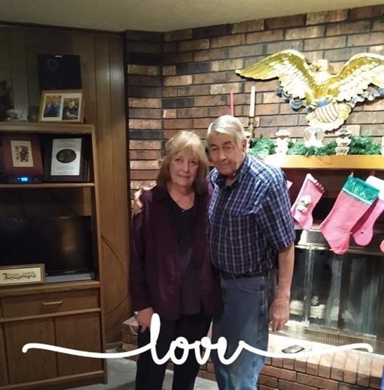 Elderly couple smiles in front of a decorated fireplace with stockings, capturing a joyful moment.