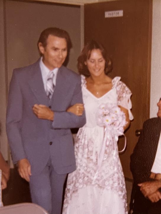 A couple dressed in formal clothing walks side by side at a festive gathering inside.