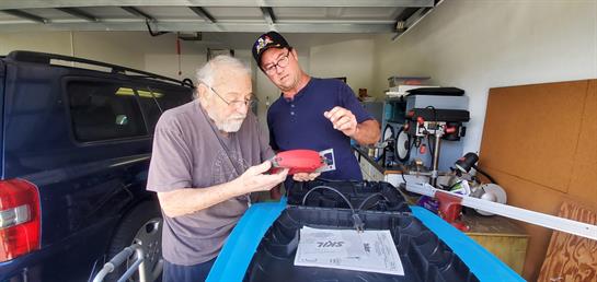 Two men examine tools and equipment in a garage, engaged in a thoughtful discussion.