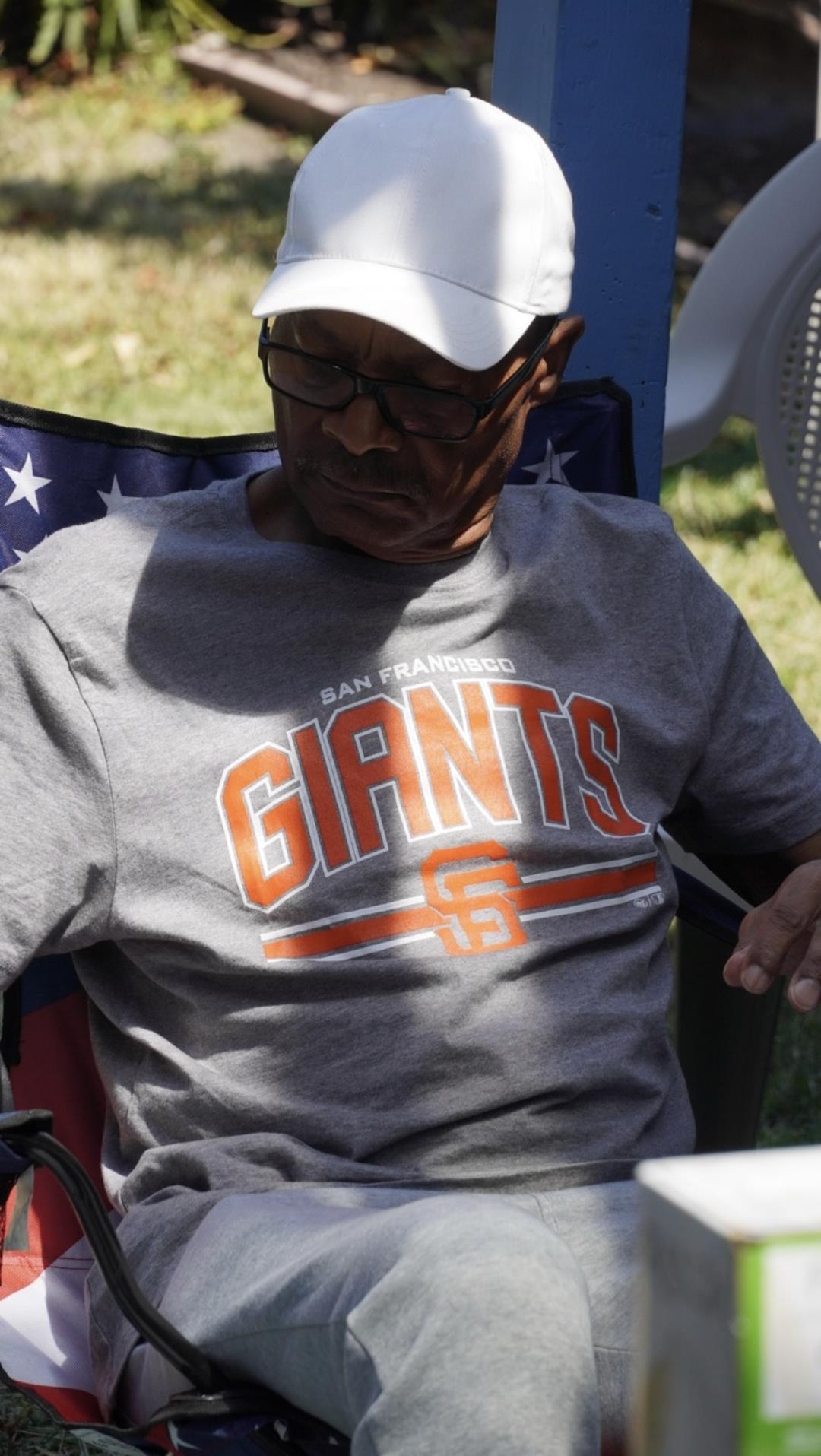 An elderly man sits comfortably in a chair, enjoying the sunshine while wearing a sports shirt.