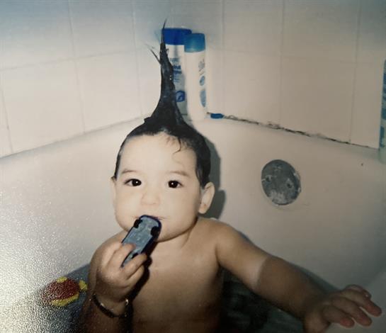 A young child with a unique hairstyle sits in a bath, holding a toy while splashing water.