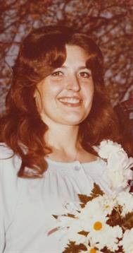 A joyful woman in a light dress and flower corsage smiles at a gathering.