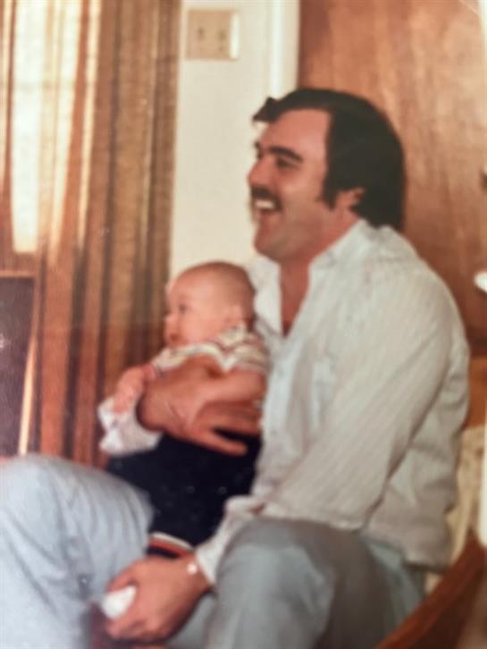 A father enjoys a playful moment with his baby, both smiling and relaxing at home.