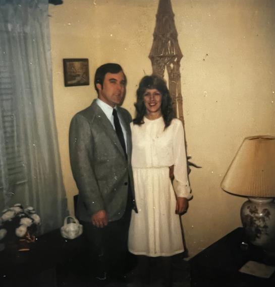 A couple in 1980s formal wear smiles in a warmly decorated living room.