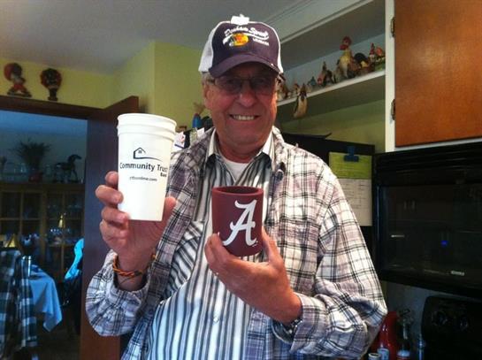 Man smiles while holding two coffee cups, showcasing his school pride in a bright kitchen.