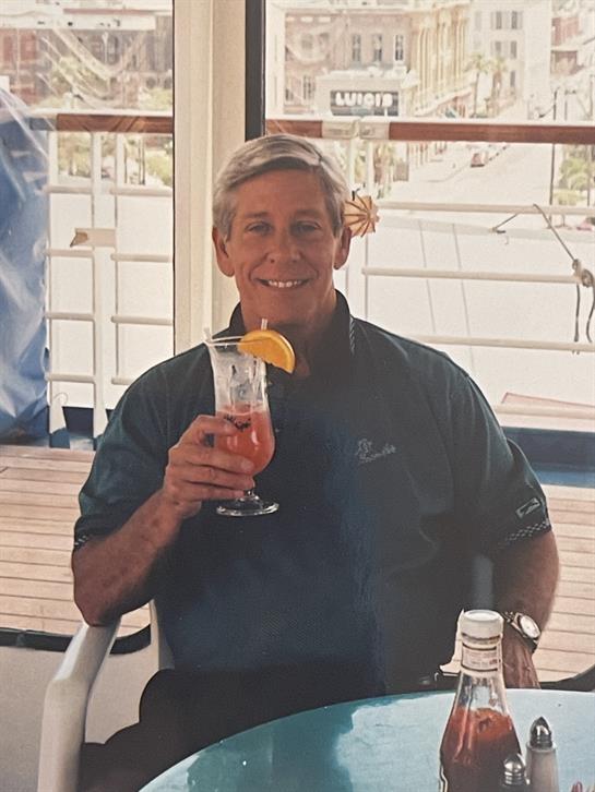 A man smiles while holding a colorful tropical drink under sunny skies on a ship deck.