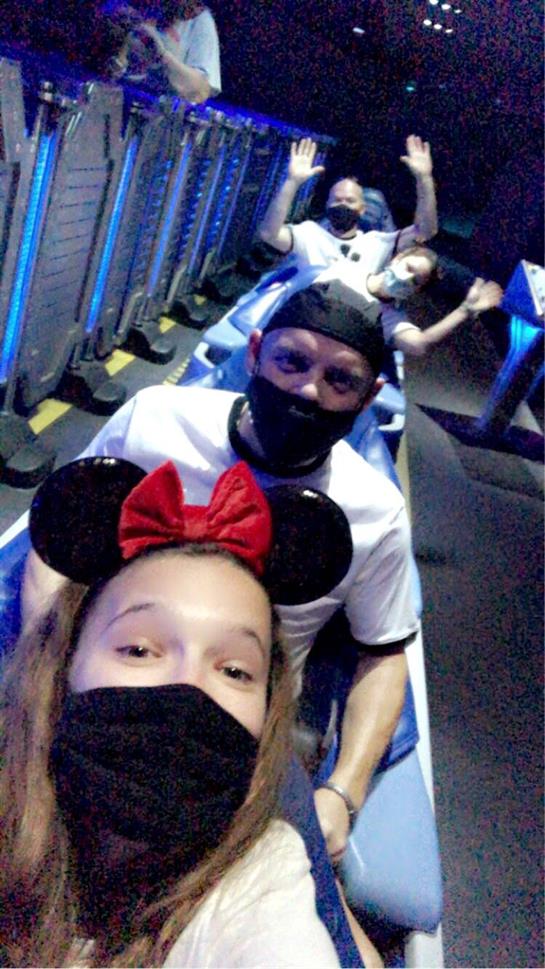 A child and an adult share excitement on a roller coaster, both wearing face masks and mouse ears.