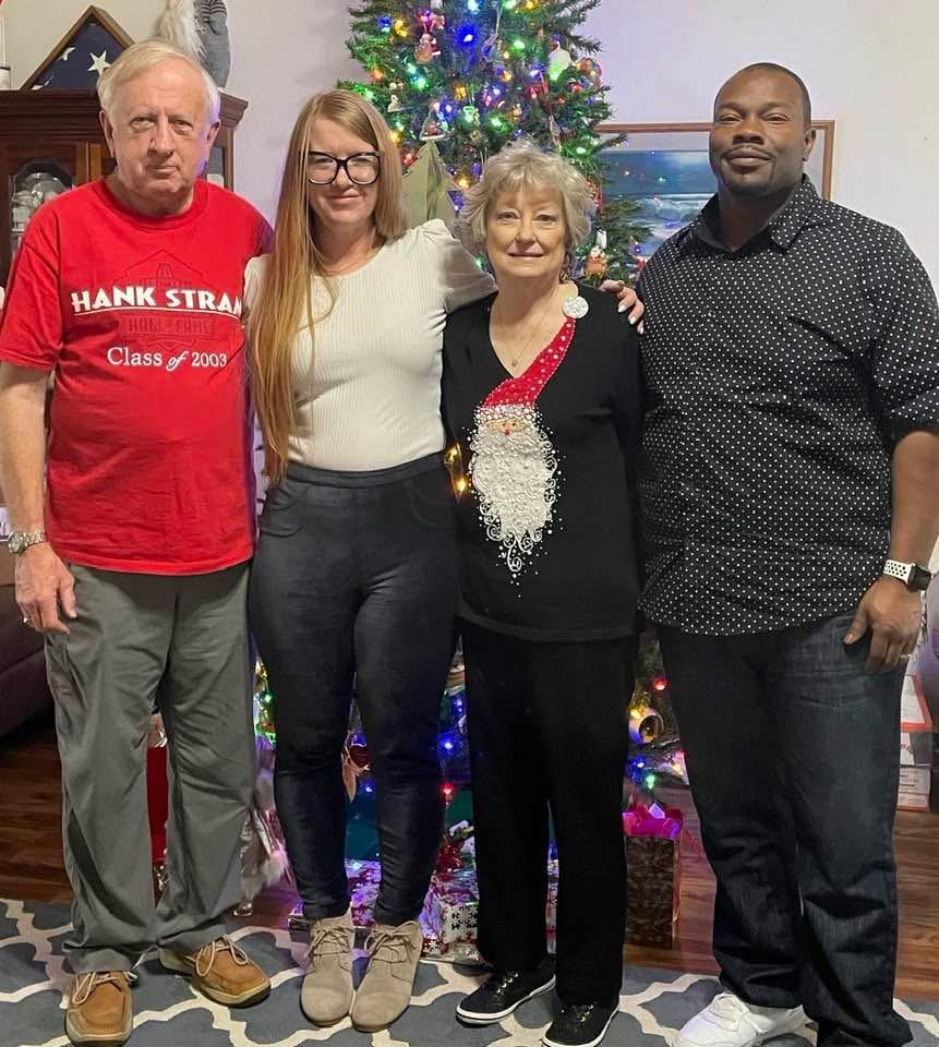 Four people stand together in a room decorated for Christmas with a tree lit up in the background.