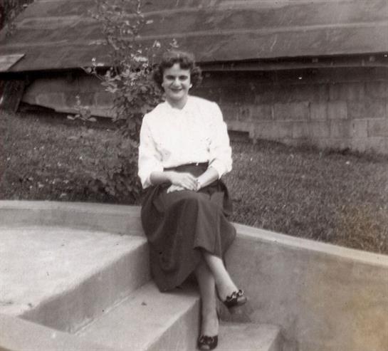 A woman poses on stone steps with a serene garden setting in the background, enjoying a sunny day.