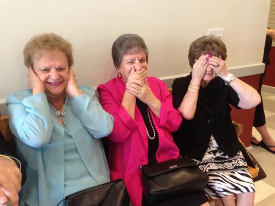 Three elderly women express joy and laughter while covering their eyes and ears during a gathering.