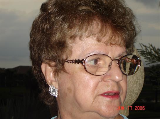 An older woman with short hair and glasses smiles warmly while seated in a comfortable space.
