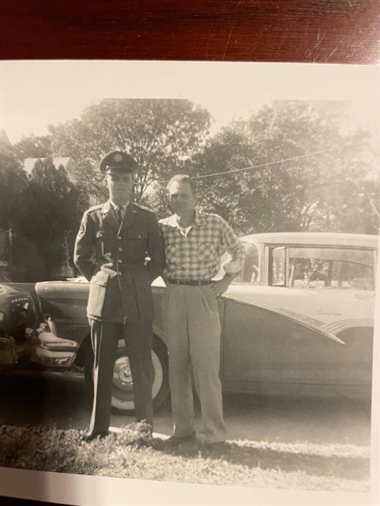 Two men stand outside: one in military gear, the other casual, by a classic car.