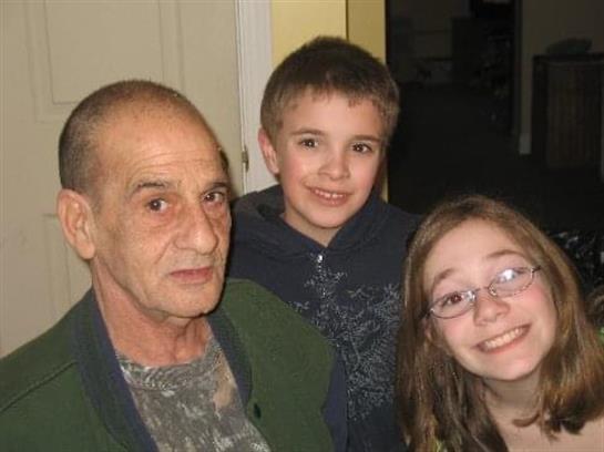 An older man poses with two children, all smiling happily in a warm indoor setting.