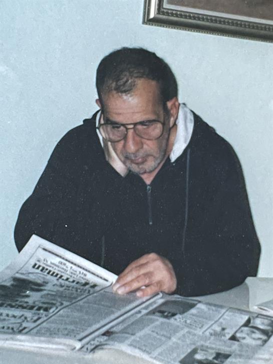 A senior man is seated at a table reading a newspaper, focused on the text and wearing glasses.