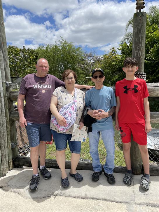A family stands together near a wooden fence, smiling under a partly cloudy sky.