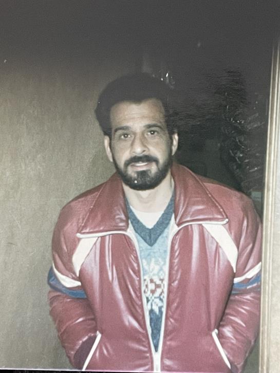 A man stands smiling inside a dimly lit room wearing a red jacket.