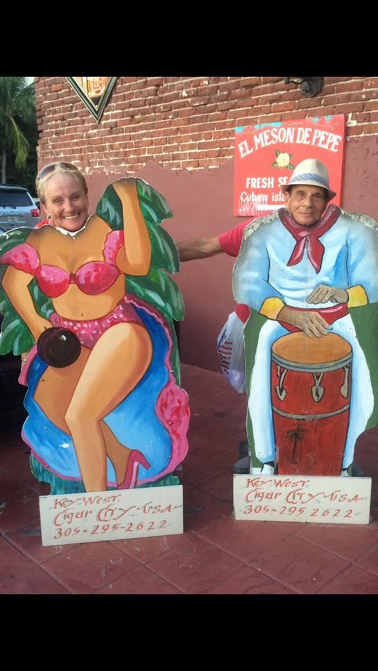 Two people pose behind vibrant cardboard cutouts at a lively summer festival by the beach.