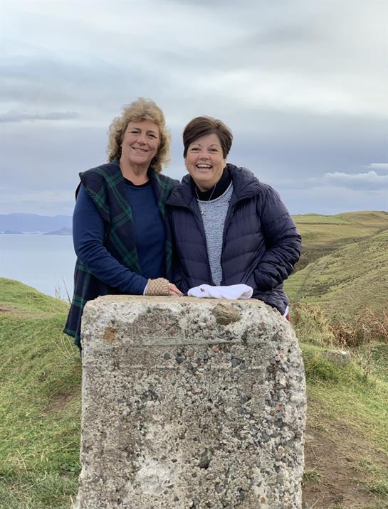 Two friends stand happily beside an ancient stone marker on a hillside, enjoying a picturesque view.