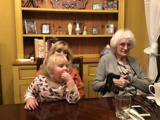 Three generations gather at a wooden table in a warm, homey atmosphere, sharing moments together.