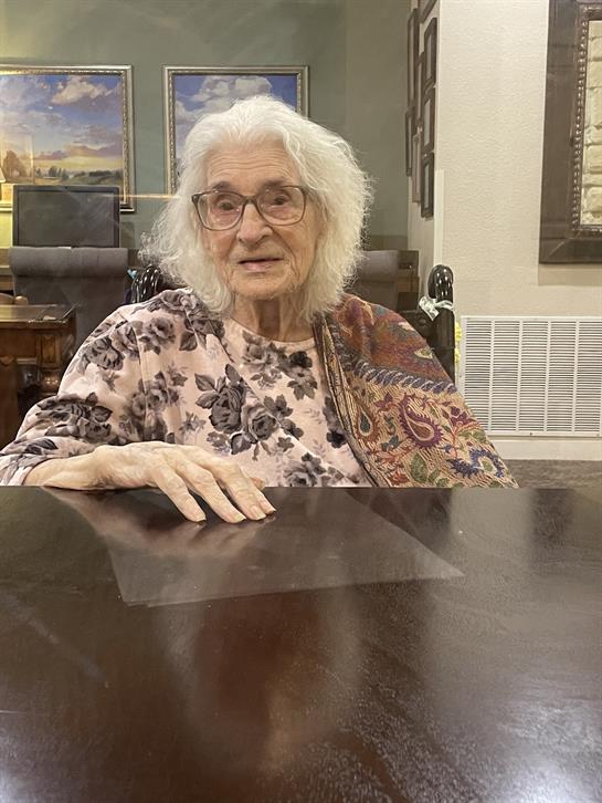 A senior woman with white hair and glasses is seated at a table, wearing a floral patterned garment.