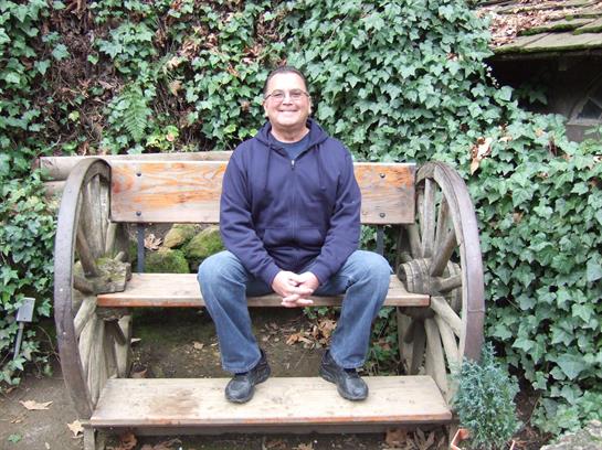 A man smiles on a wooden bench amid lush greenery in a peaceful outdoor setting.
