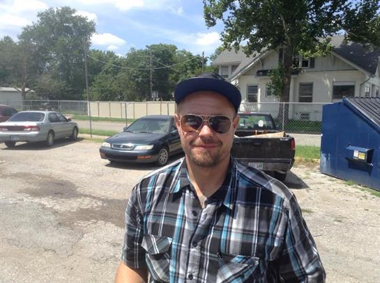 A man with sunglasses and a plaid shirt poses for the camera in a parking lot under a clear sky.