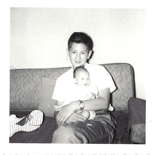 A boy cradles a baby in his arms, both sharing a moment on a couch in a cozy living room.