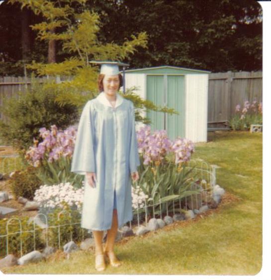 A young woman in a blue graduation gown stands barefoot in a vibrant flower garden.