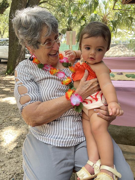 An elderly woman smiles as she holds a baby girl, both enjoying a sunny day in the park.
