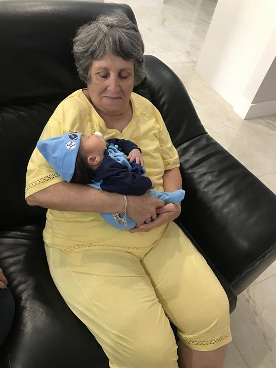A grandmother sits on a black chair, tenderly holding her sleeping grandchild in a cozy outfit.