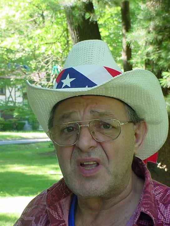 Older man talks with a serious expression while wearing a festive cowboy hat outdoors.