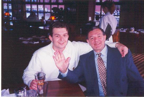 Two men are seated at a table in a busy restaurant, smiling and waving.