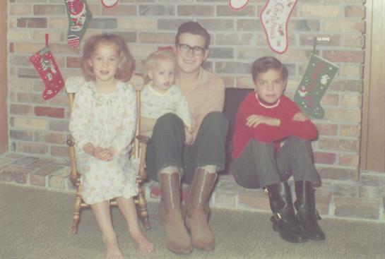 Four children, one in a rocking chair, smile together by a decorated fireplace during winter.