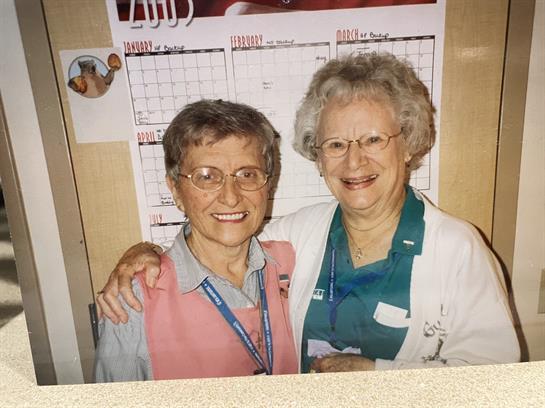 Two cheerful elderly women embrace and smile in a community center, showcasing their friendship.