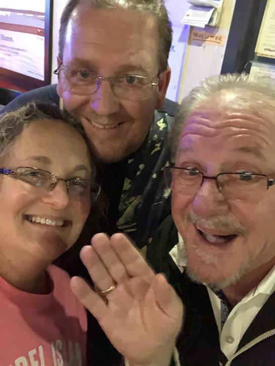 Three friends smile and wave at the camera during a joyful evening gathering.