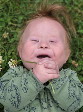 A joyful child lies in a meadow, smiling and holding a plant, enjoying nature's beauty.