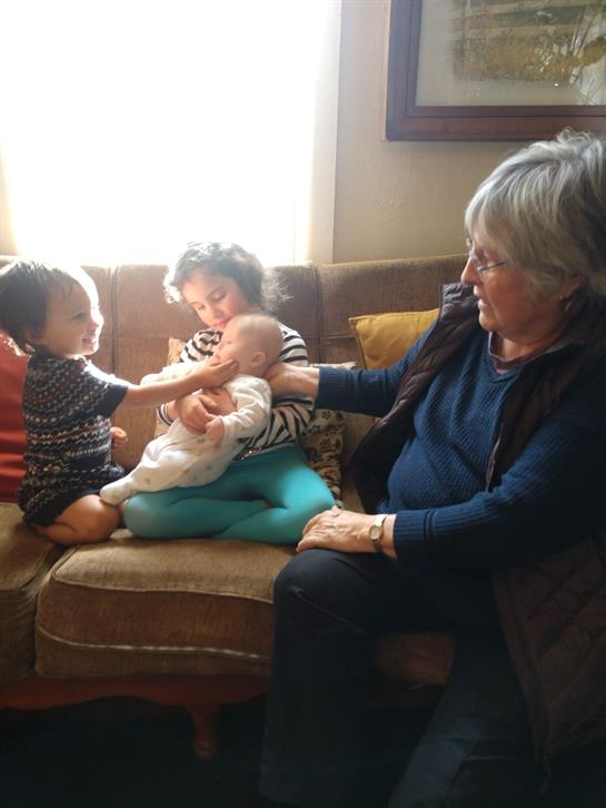 Children interact playfully with their grandmother on a cozy couch in the sunlight.