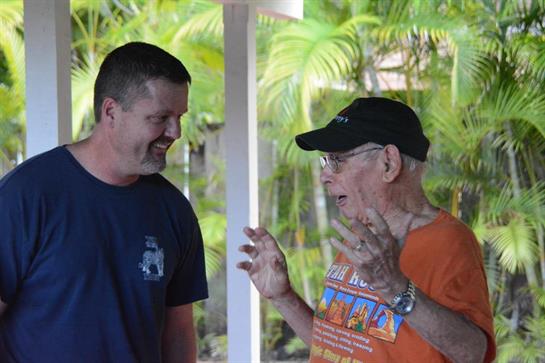 Two men talk enthusiastically under a pavilion while tropical plants surround them.