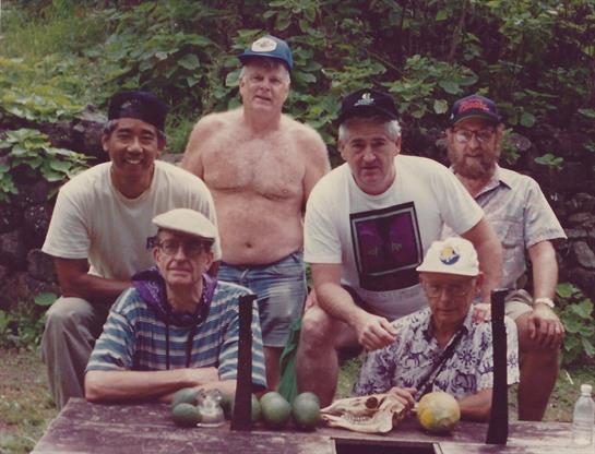 A group of older men in hats gather outdoors around a table of fruits, enjoying each other.