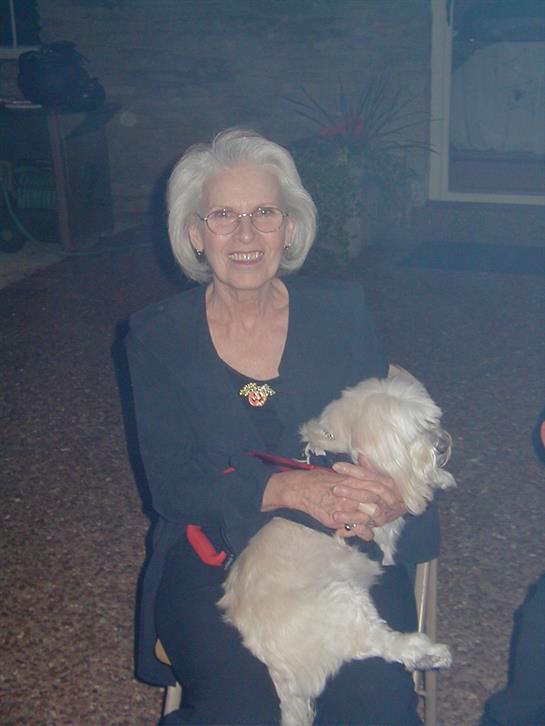 An elderly woman smiles joyfully, holding her small dog at a warm indoor gathering.