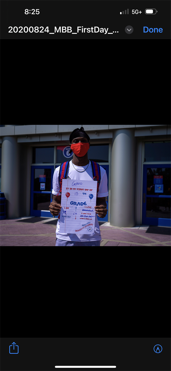 Student proudly holds a sign marking the first day of school while wearing a mask at the entrance.
