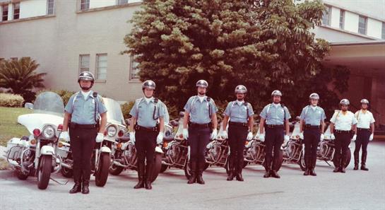 Officers in uniform line up with motorcycles near a building on a sunny day.