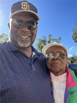 A man and elderly woman smile at the camera on a sunny day, surrounded by greenery.