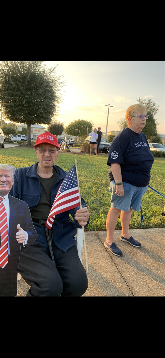 Elderly man sits with a Trump cutout and flag while a woman stands nearby during a rally.