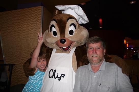 A cheerful girl gives a peace sign next to Chip, the mascot, and an older man at a festive event.