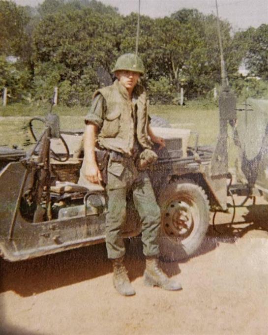 A soldier poses beside a military jeep while participating in a training exercise outdoors.