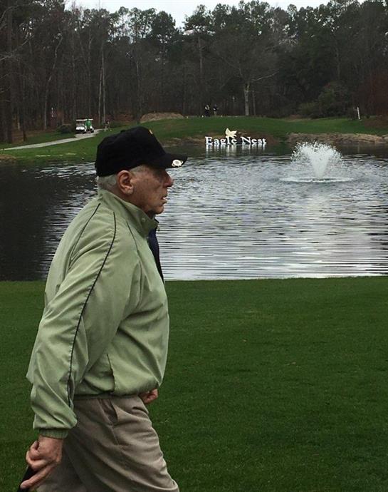 An elderly man strolls past a pond on a golf course, enjoying the calm surroundings and nature.