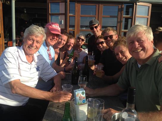 Group of friends sharing laughs and drinks at an outdoor table under a clear blue sky.