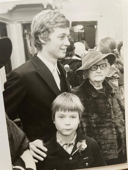 Young man stands with an elderly woman and a boy, all dressed formally at a crowded event.