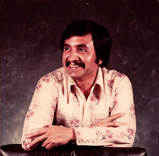 A man with a mustache smiles joyfully while leaning on a table, dressed in a floral-patterned shirt.